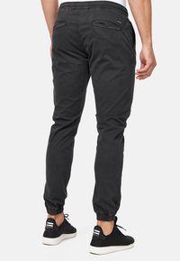 Homme portant un pantalon de jogging noir avec des poignets élastiques et des baskets noires, vu de dos sur un fond blanc uni.