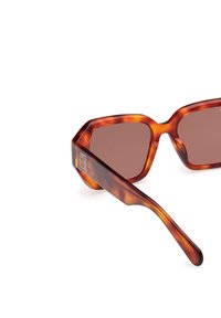 Ochelari de soare tortoiseshell maro cu un cadru geometric, având lentile maro închis și un detaliu cu logo pe tampla.