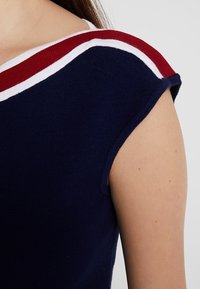 Hombro y parte superior del brazo vistiendo una camiseta de punto azul marino con rayas rojas y blancas a lo largo del escote.