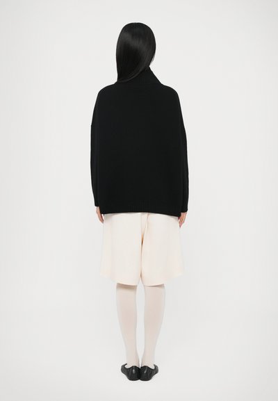 Pull en maille noire avec un col montant, associé à un short beige clair. Le mannequin porte des collants neutres et des ballerines noires, vu de dos.