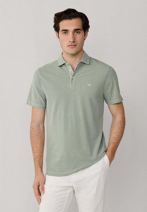 Jonge man die een lichtgroen poloshirt met korte mouwen en witte broek draagt, staand tegen een effen lichte achtergrond met één hand in zijn zak.