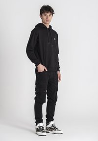 Sudadera negra con capucha y bolsillo delantero, combinada con pantalones cargo negros que tienen múltiples bolsillos y zapatillas altas de diseño.
