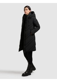 Sort pudded coat med hætte, med diagonalt quiltet design, lynlås foran og sidelommer. Båret over sorte bukser og støvler.