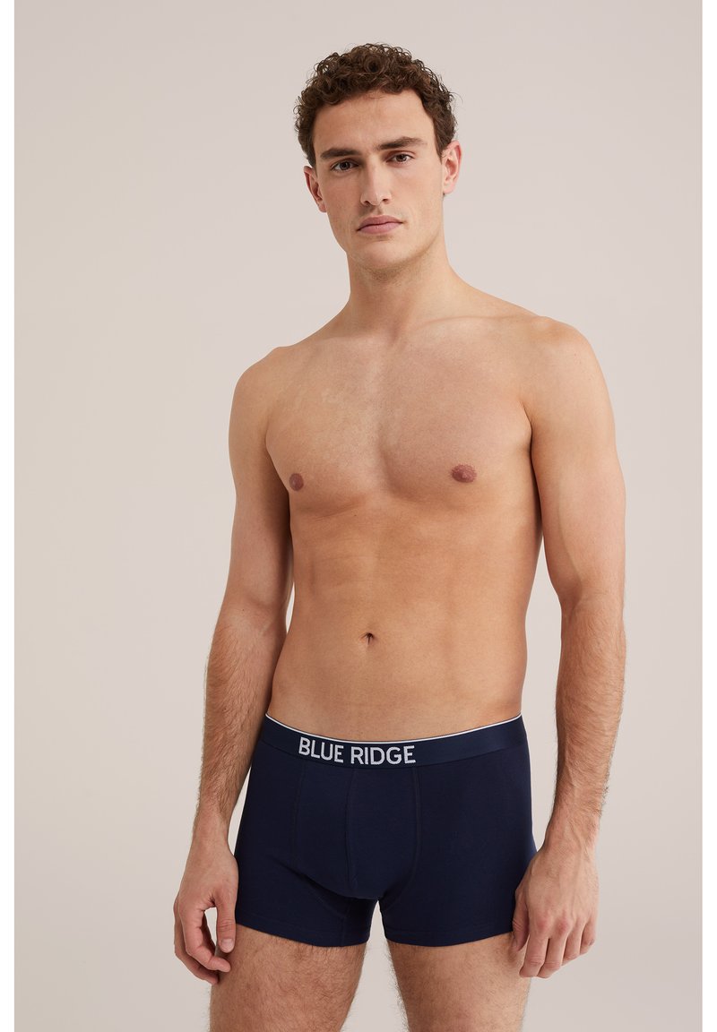 Jeune homme torse nu portant un boxer bleu marine Blue Ridge sur un fond clair uni.