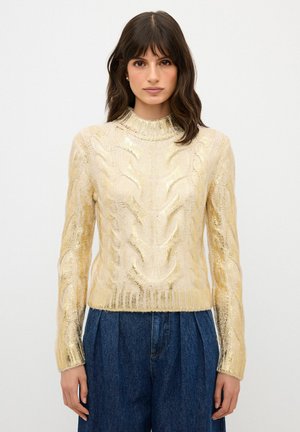 Maglione lavorato a maglia in morbido filato dorato con un motivo a treccia testurizzato, collo alto e orlo a coste; abbinato a pantaloni blu a vita alta.