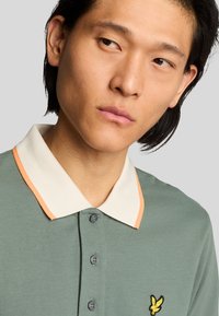 Grön polotröja med strukturerad yta, med en vit krage med orange kant och en liten broderad logotyp på vänster bröst.