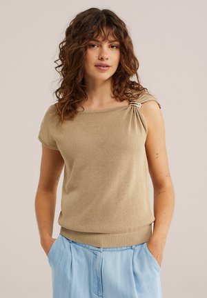 Vrouw met krullend haar die een beige top met korte mouwen en een gespdetail op de schouder draagt, en lichtblauwe geplooide broek, handen in de zakken.