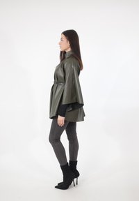 Olijfgroene nep leren cape-jack met hoge kraag, een taille met riem en soepel vallende mouwen, gecombineerd met grijze strakke broek en zwarte stiletto laarzen.