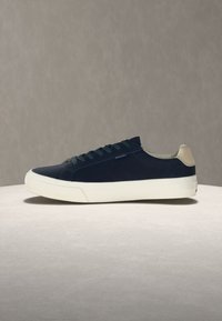 Σκούρο μπλε παπούτσι χαμηλού τύπου με λευκή λαστιχένια σόλα και μπεζ ταμπ πουκάμισο στην φτέρνα, παρουσιάζεται σε ελαφρύ στρογγυλό επιφάνεια πάνω σε ουδέτερο φόντο.