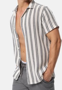 Mann in einem kurzärmligen weißen Hemd mit schwarzen vertikalen Streifen, offen getragen, mit grauen Hosen und einer Hand in der Tasche.