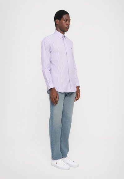 Šviesiai violetinė ir balta dryžuota marškinėliai su sagomis ir apykakle, derinami su šviesiai mėlynais tiesiais džinsais ir baltais sportbačiais.