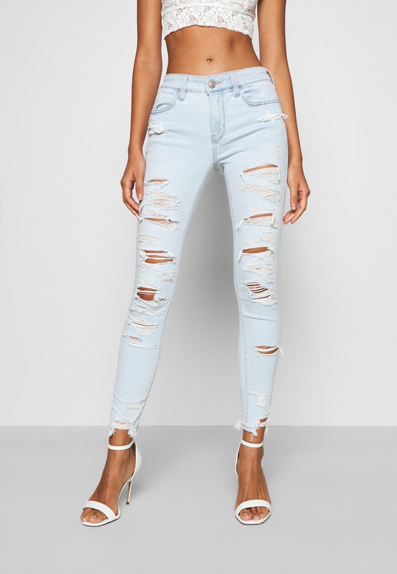 Light blue denim skinny jeans with distressed rips, high waist, and frayed hem; paired with white heeled sandals.