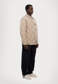 Giacca trapuntata beige con colletto, tasca sul petto singola e bottoni; abbinata a pantaloni neri e sneakers crema. Modella in piedi di lato.