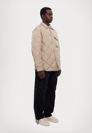 Béžová prešívaná bunda s golierom, jednou náprsnév kapsou a gombíkmi; kombinovaná s čiernymi nohavicami a krémovými teniskami. Model stojí zboku.