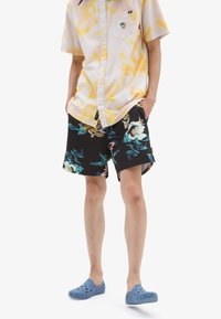 Camicia a maniche corte con bottoni in tie-dye giallo, pantaloni corti neri a fiori con accenti blu e rosa, e zoccoli blu perforati.