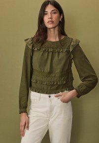 Zielona oliwkowa bluzka z falbankami na ramionach, teksturowym detalem przy dekolcie i warstwowymi falbankami wzdłuż dolnej krawędzi. W połączeniu z białymi spodniami.