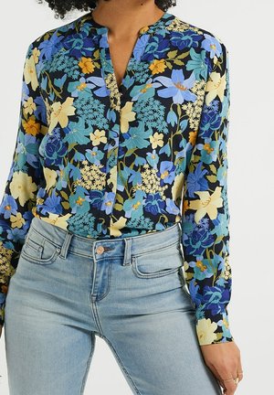 Kobieta ubrana w niebieską, żółtą i zieloną kwiecistą koszulę na guziki, włożoną do jasnoniebieskich dżinsów, na tle jednolitego białego tła.