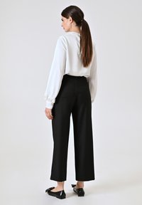 Blusa bianca a maniche lunghe con collo alto e chiusura posteriore, abbinata a pantaloni neri a gamba larga a vita alta. Scarpe flat nere con fiocchi.