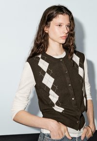 Young woman with long wavy hair wearing a brown argyle sweater vest over a white long-sleeve shirt with hands in jeans pockets.
