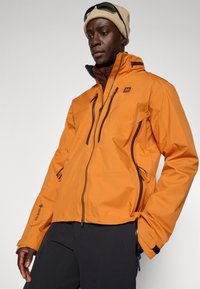 Waterdichte oranje jas met zwarte ritsen, ritssluitingen, hoge kraag en een logo op de borst. Heeft een getextureerd oppervlak.