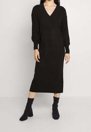 Vrouw draagt een zwarte geribbelde midi-jurk van gebreide stof met lange mouwen en V-hals, gecombineerd met zwarte enkellaarsjes met een middelhoog hakje, op een witte achtergrond.