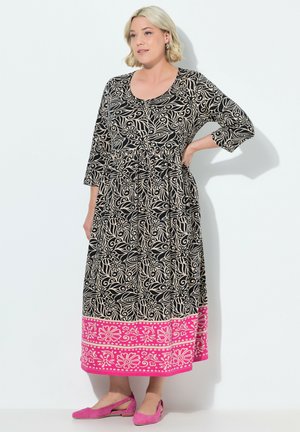 Robe longue noire et beige à motifs floraux avec une bordure rose à motifs, manches 3/4 et taille cintrée. Portée avec des chaussures roses.