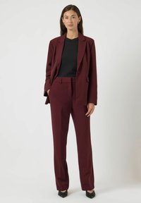 Kobieta stoi, ubrana w bordową marynarkę i spodnie, czarną bluzkę oraz czarne buty w szpic, na tle jasnego, jednolitego tła.