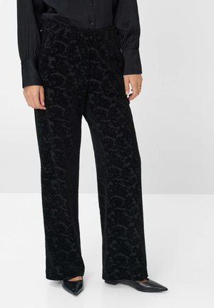 Woman wearing black patterned wide-leg velvet trousers, paired with black pointed-toe flats and a black buttoned blouse.