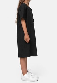 Jeune personne debout de profil portant une robe noire ample longueur genoux et des baskets blanches sur un fond uni.
