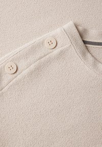 Maglione beige con texture e colletto a coste, dotato di due bottoni color crema sulla spalla e un dettaglio a righe bianche e nere all'interno del colletto.