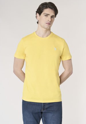 Uomo con capelli scuri che indossa una T-shirt gialla a maniche corte semplice e jeans blu, in piedi con le mani dietro la schiena davanti a uno sfondo semplice.