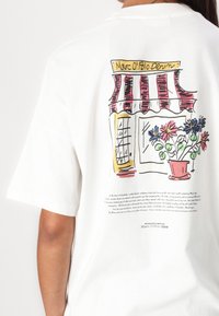 White cotton t-shirt featuring a colorful illustrated storefront with red awning and potted flowers on the back, with text below the design.