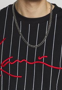 Czarna koszula z krótkim rękawem z białymi pionowymi paskami, z czerwoną haftowaną czcionką. Na szyi widoczny łańcuch naszyjnika.