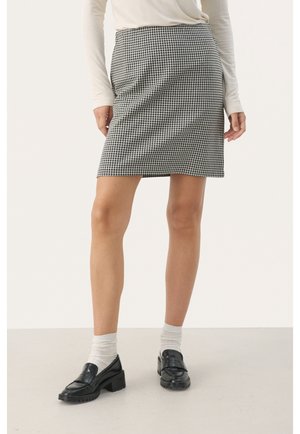 Femme portant une jupe pied-de-poule noire et blanche, un haut à manches longues blanc, des chaussettes blanches et des mocassins noirs épais, debout sur un fond uni.