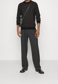 Maglione nero a trama lavorata a maglia con orlo a coste scuro, abbinato a pantaloni grigi larghi e scarpe in pelle nere, oltre a una piccola borsa a tracolla.