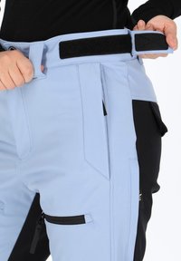 Light blue pants with black accents, featuring a Velcro waistband, side pockets, and zippered thigh pocket. Smooth texture and tailored fit.