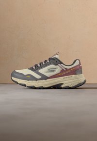 Baskets athlétiques beige et grises avec des accents bordeaux, tige en mesh, semelle épaisse mouchetée et marquage "Gotrail" sur le côté.