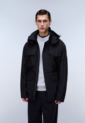 Young man with slicked-back hair wearing a black hooded jacket over a light gray sweater, standing against a plain light background.