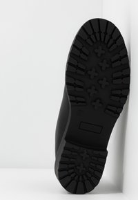 Semelle d'une botte noire avec des motifs de bande de roulement profonds, y compris des formes en croix et des rainures, présentée sur un fond blanc.