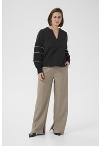 Schwarze Bluse mit V-Ausschnitt und puffärmeln sowie weißen Stickdetails, kombiniert mit beigen, weit geschnittenen Hosen mit seitlichen Schlitzen und schwarzen Schuhen.