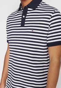 Dospělý ve svetříku s krátkým rukávem, pruhovaném modro-bílém, s modrým límečkem a dvěma bílými knoflíky.