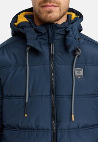 Giacca puffer navy con cappuccio, zip grigie e dettagli testurizzati. Cappuccio interno giallo. Presenta un logo sulla parte del petto.