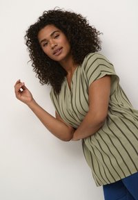 Blouse vert olive clair avec des rayures verticales foncées, manches courtes, col en V avec des boutons, et coupe décontractée, portée sur un jean bleu.