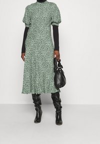 Blommig grön midi-klänning med korta puffärmar, texturerat tyg och utsvängd kjol, parat med en åtsittande svart topp och svarta knähöga stövlar.