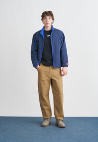 Μπλε μπουφάν με μπλε διακοσμητικά, μαύρο t-shirt, μπεζ παντελόνι και ανοιχτό γκρι μπότες. Το μοντέλο στέκεται μπροστά σε ουδέτερο φόντο πάνω σε μπλε δάπεδο.