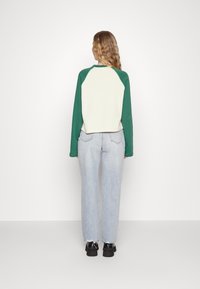 Sweatshirt raglan de manga longa verde e creme com a bainha arredondada, combinado com jeans azul claro e calçado preto, visto de costas.