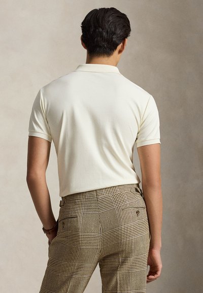 Muž stojící zády, v krátkém bílém polotriku a béžových károvaných kalhotách s rukama v kapsách na jednobarevném pozadí.