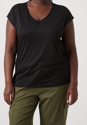 Person trägt ein schwarzes Kurzarm-V-Ausschnitt-Shirt und olivgrüne Hosen mit Seitentaschen, steht vor einem schlichten hellen Hintergrund.