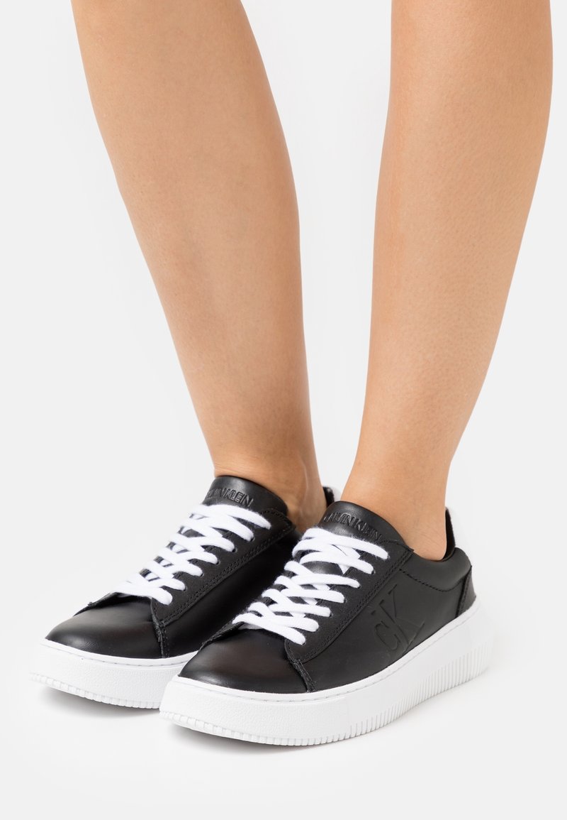 Les baskets en cuir noir comportent des lacets blancs, un bout rond et une épaisse semelle en caoutchouc blanche à motifs rainurés. Logo embossé sur le côté.