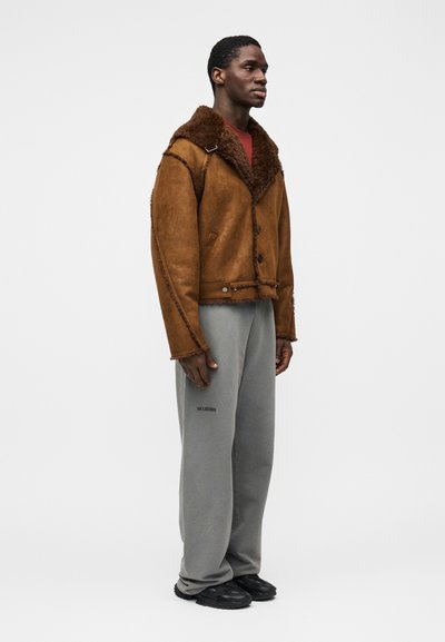 Καφέ μπουφάν shearling με κουκούλα, που διαθέτει υφή και κλείσιμο με κουμπιά. Συνδυασμένο με γκρι φόρμες και μαύρα αθλητικά παπούτσια.